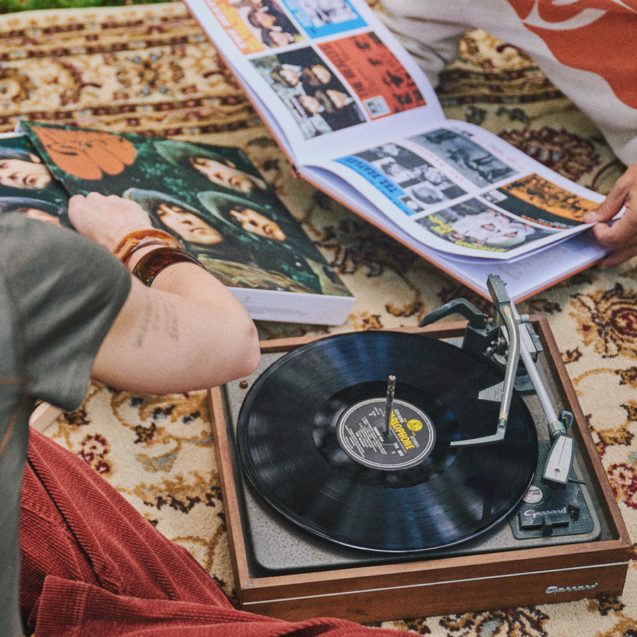 A wooden turntable with a black vinyl record is set on a patterned rug, surrounded by people holding open books or magazines in a cozy indoor setting. Showing the 5LP Box Set Exclusive items