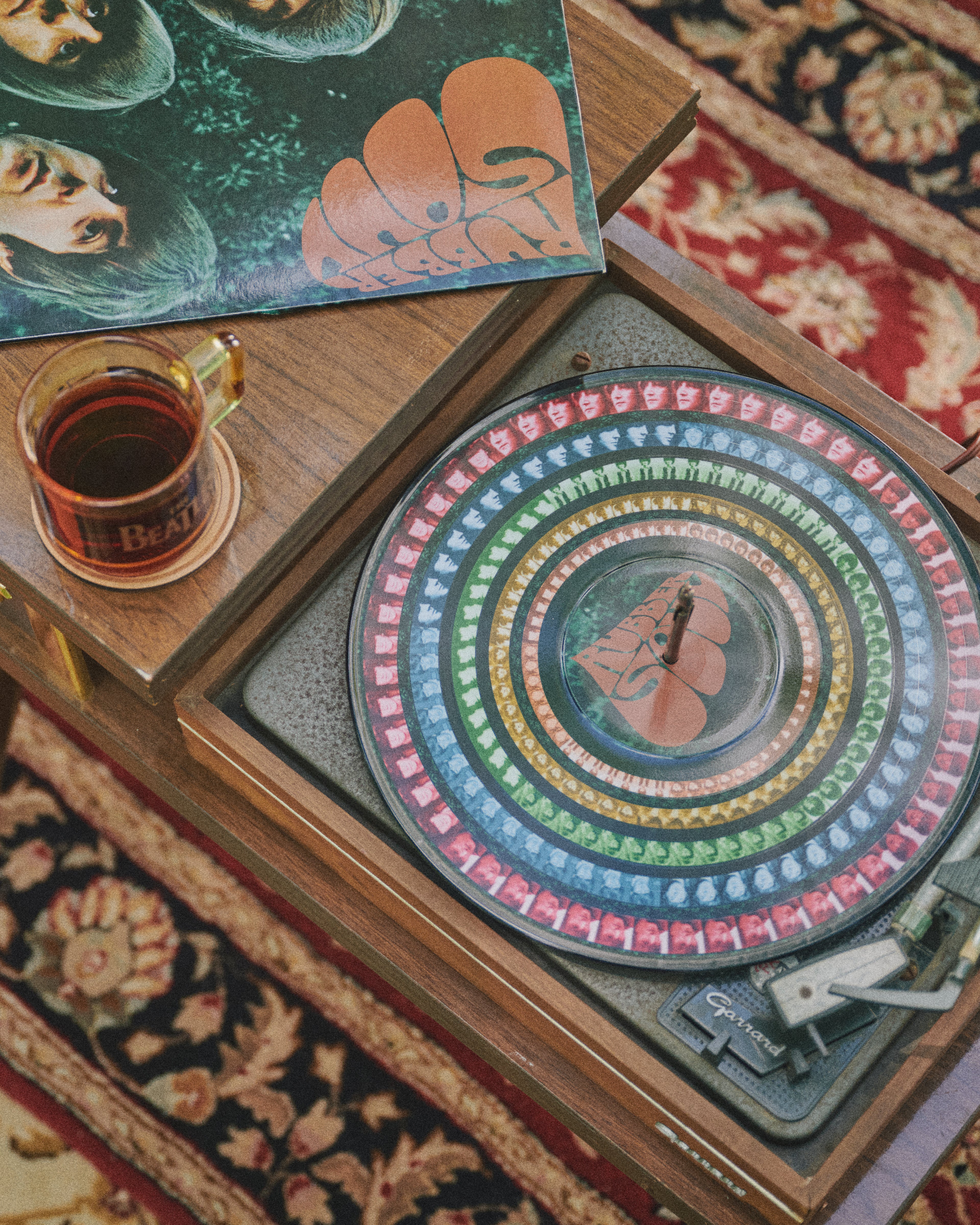 A vintage wooden turntable with a colorful concentric-patterned vinyl record, styled on a patterned rug with a glass of tea and album art nearby. Rubber Soul Zoetrope
