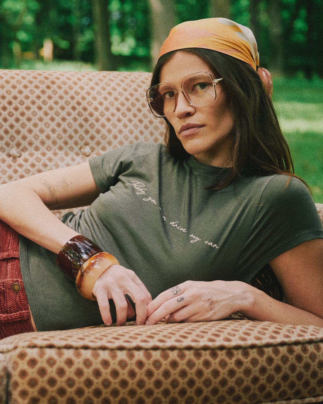 A woman wearing the Rubber Soul Baby You Can Drive My Car Handwritten Baby Tee, a mustard yellow headscarf, and glasses, reclining on a brown patterned armchair in an outdoor setting