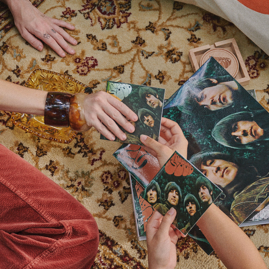 Several people's hands are gathered around the 4CD Box Set Exclusive, placed on a patterned beige rug with a glass bowl and rust-colored fabric nearby.