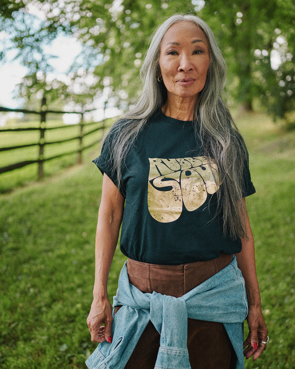 A woman with long gray hair stands outdoors in a grassy field wearing Rubber Soul Black Gold Foil Tee with a gold elephant design, brown pants, and a light blue shirt tied around her waist.