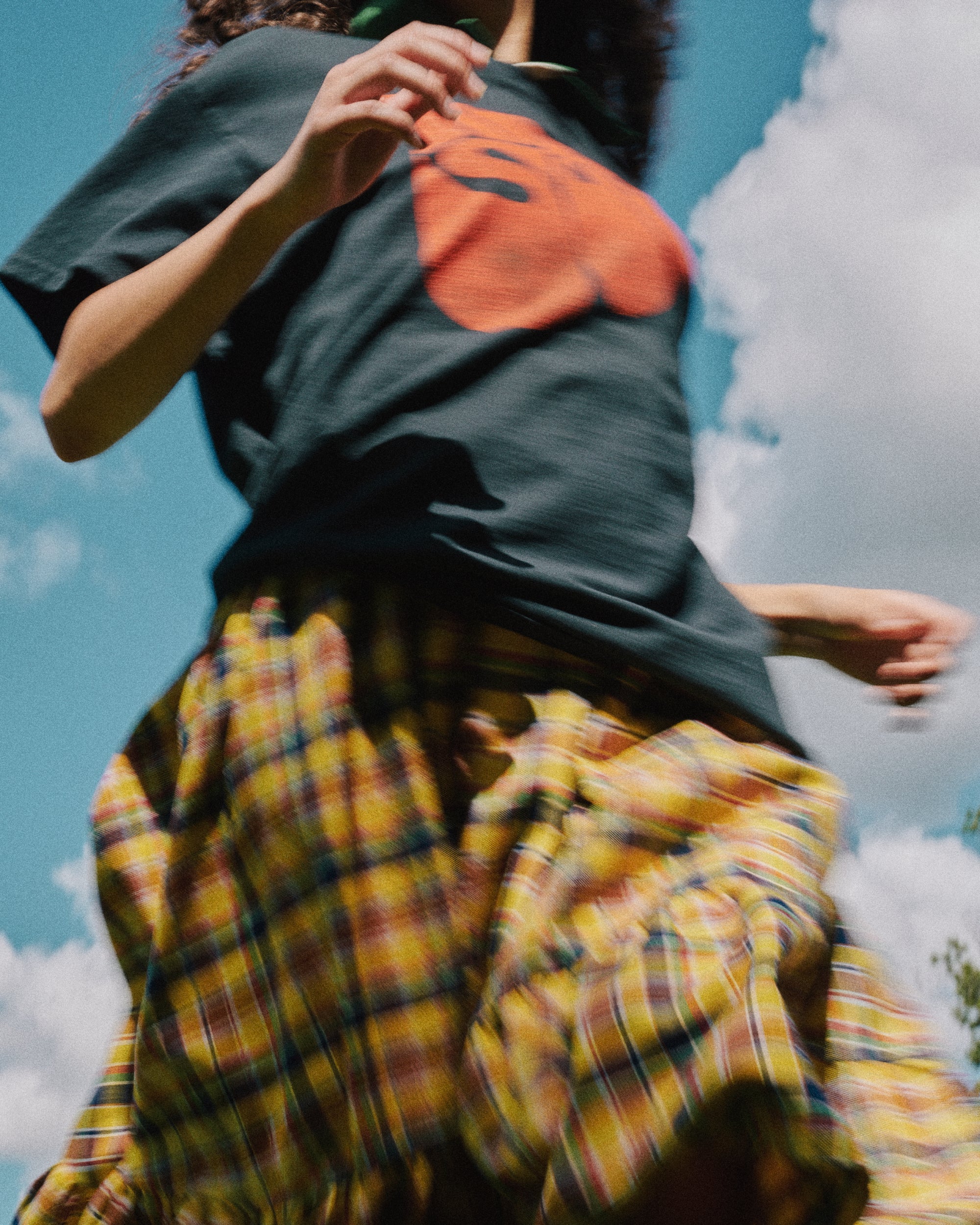 A person outdoors wearing Rubber Soul Black & Orange Distressed Tee and a yellow, red, and blue plaid skirt, captured in motion against a blue sky with clouds.