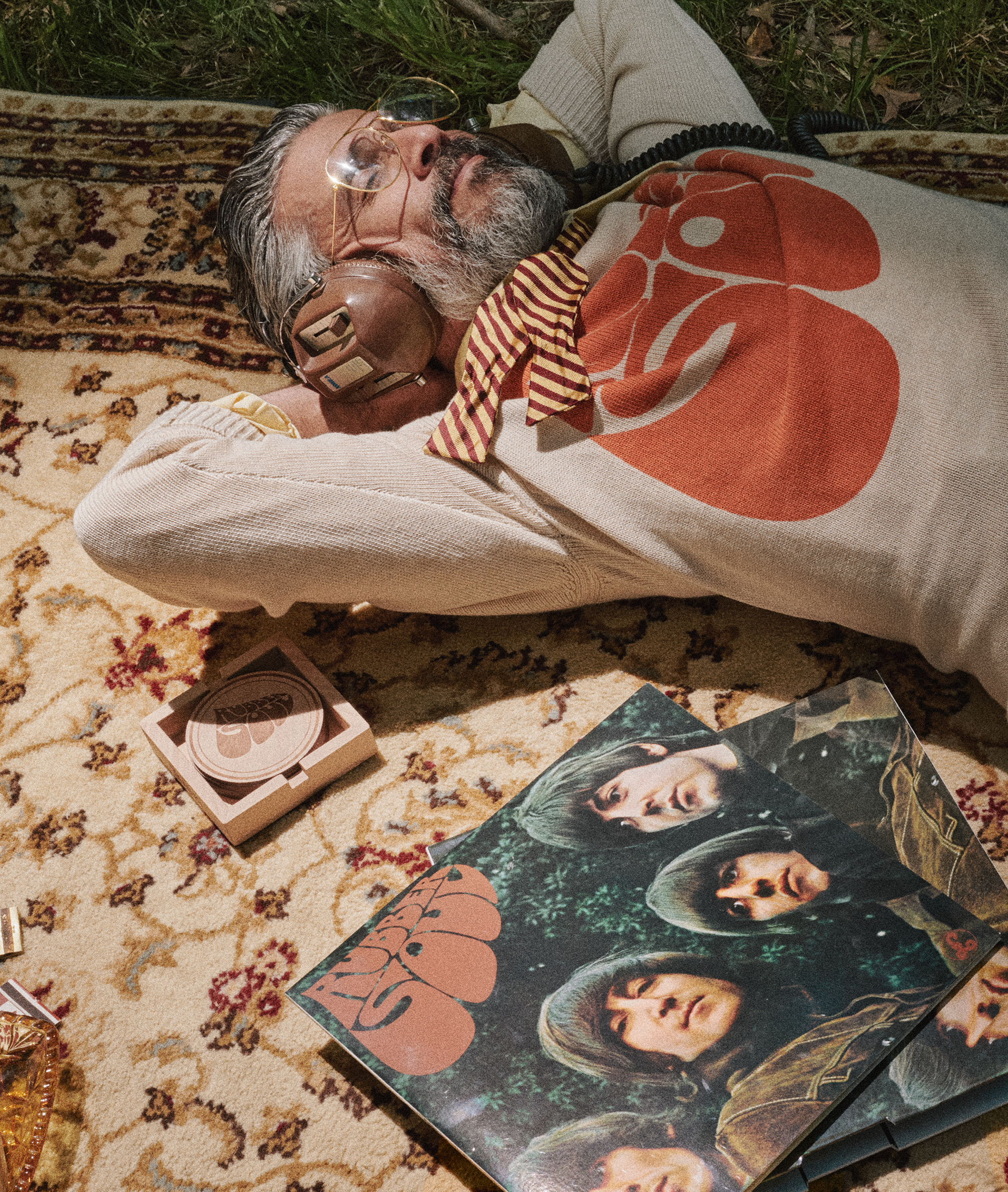 A man lies on a patterned rug outdoors, wearing a cream knit sweater with a large orange Rubber Soul graphic and a striped neck scarf. A Rubber Soul record sleeve and a small round case are placed nearby on the rug.
