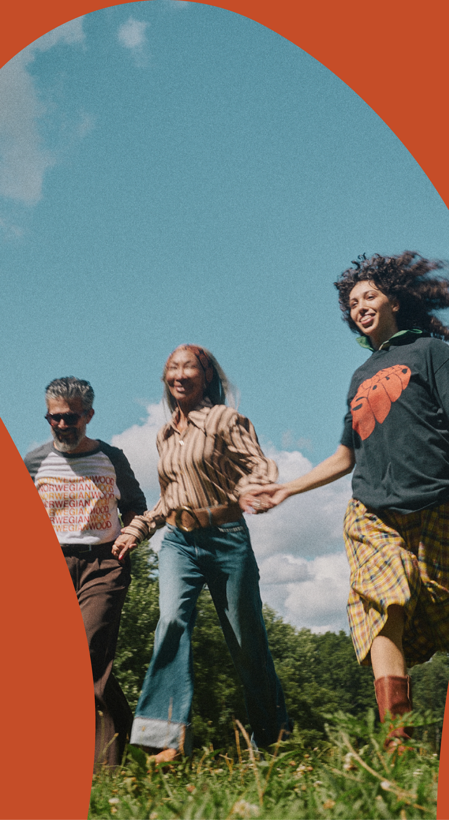 Three people holding hands and smiling outdoors in a grassy field under a blue sky, framed by a bold orange arch graphic on the left and right edges. People are wearing the Rubber Soul Norwegian Wood Raglan, Phix X The Beatles Brown Stripe Point Collar Shirt, and the Rubber Soul Black & Orange Distressed Tee.